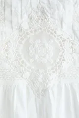 Blusa blanca de algodón con cuello redondo ribeteado en encaje y mangas cortas holgadas. Presenta un canesú fruncido con jaretas y detalles de broderie en la parte frontal.