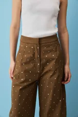 Pantalón marrón de corte ancho, tiro alto y diseño holgado, adornado con pequeños apliques metálicos distribuidos por toda la prenda.