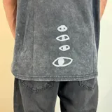 Remera de corte oversize con efecto de lavado ácido en color negro. Presenta un estampado rectangular central en tonos violeta con la palabra 'VISIONS' y una escena de surfistas en el mar.