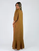 Vestido largo de lino color marrón tostado, con mangas 3/4 abullonadas, escote en V con volados y botones frontales. Cuenta con bolsillos laterales y detalle de volado en el dobladillo inferior.
