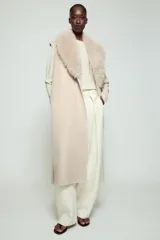 Chaleco largo de mezcla de lana color beige, de corte recto con manga sisa. Cuenta con cuello solapa con aplicación de pelo sintético extraíble, bolsillos laterales ocultos y un cinturón ajustable del mismo tejido para anudar en la cintura.