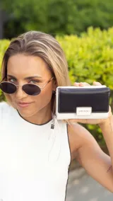 Billetera mediana de diseño bicolor, combinando una solapa en tono dorado metalizado con cuerpo en color negro. Presenta cierre perimetral, correa de muñeca extraíble con detalle de borla y placa metálica con logo de la marca en el frente.