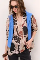 Blusa de manga larga con estampado abstracto de hojas en tonos beige, marrón y negro, con cuello en V y cierre frontal con botones ocultos.