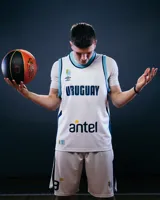 Camiseta de básquet Umbro para hombre, modelo alternativo de la selección de Uruguay. Color blanco con ribetes celestes en cuello y sisas. Presenta el nombre "URUGUAY" en azul oscuro en el pecho, el logo de Umbro, el escudo de la selección y el sponsor "antel" en amarillo y azul.