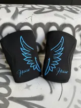 Par de rodilleras negras de neopreno de 7mm con logo de alas y nombre de la marca en color celeste.