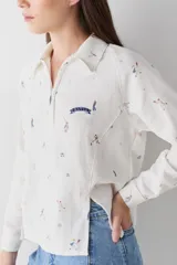 Camisa de manga larga color blanco con cuello polo y cierre frontal parcial. Presenta un estampado integral con motivos deportivos y un detalle bordado en el pecho.