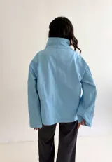 Campera celeste de algodón con diseño versátil de cuello alto envolvente, cierre frontal cruzado con botones metálicos a presión y corte boxy.