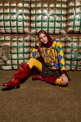 Sweater tejido de algodón con cuello alto marrón, diseño con rombos amarillos, verdes y azules, y bordado frontal de dos figuras.