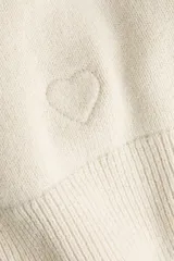 Saco entallado de punto fino en color beige, con cuello redondo, cierre frontal de botones y detalle de corazón bordado en el frente. Presenta terminaciones acanaladas en cuello, puños y bajo.