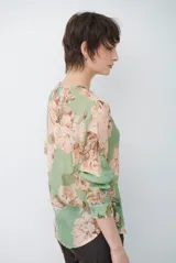 Camisa de manga larga con fondo verde y estampado floral en tonos rosados y beige, cuello redondo con pequeña abertura en V y cierre frontal con botones.