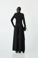 Pantalón negro de tiro bajo, corte ancho y recto, con un detalle de tela superpuesta en forma de bolado o drapeado asimétrico en la parte frontal de una de las piernas.