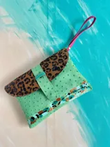 Sobre de mano confeccionado en cuero verde texturado, con solapa de cuero con estampado animal print tipo leopardo, tira de cierre con broche imán y detalles pintados a mano. Incluye una correa de muñeca en color fucsia.