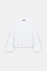 Camisa blanca de cuello subido con detalle de volantes en el frente y el cuello, manga larga abullonada con puños elásticos y cierre frontal con botones.