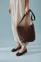 Cartera tipo morral de cuero color beige, con diseño trenzado artesanal, correa larga regulable y cierre superior.