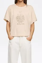 Camiseta de corte boxy color beige claro, con escote caja y manga corta con pliegues en los hombros. Presenta un estampado botánico en tonos sepia con la inscripción "Bimba y Lola".