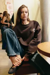 Sudadera de cuello redondo y manga larga con hombro caído, confeccionada en mezcla de algodón. Presenta un diseño liso en color marrón oscuro con un pequeño estampado gráfico a contraste en el pecho y detalle en el puño.