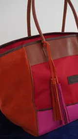 Bolso de fin de semana tipo tote, confeccionado en una combinación de lona y gamuza en tonos rojo, fucsia y marrón. Cuenta con doble asa de hombro, cierre superior y detalle de borlas colgantes en el lateral.