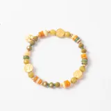 Pulsera elástica compuesta por cuentas de piedra en tonos verdes y naranjas, intercaladas con pequeñas esferas doradas y dijes circulares dorados con diseño de árbol de la vida grabado. Incluye un dije colgante adicional con la inscripción MMA.