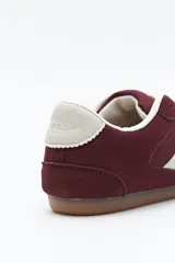 Zapatilla urbana de perfil bajo con diseño retro, confeccionada en poliuretano con detalles en textura tipo gamuza. Presenta puntera redondeada, cordones clásicos y suela de goma color caramelo.