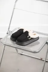 Zueco de gamuza color negro con interior forrado en piel sintética color crema. Presenta una tira decorativa con hebilla metálica dorada en el empeine y suela de plataforma plana de goma negra.