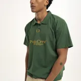 Remera tipo polo de manga corta, color verde oscuro, con cuello y abertura frontal. Presenta un estampado dorado en el pecho con el texto "PulauCrew" y un pequeño logo de corona de laurel bordado.