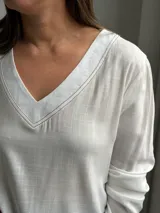 Blusa blanca de corte holgado con escote en V, mangas largas y costuras visibles en el cuello y el ruedo.