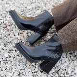 Bota corta de cuero negro con diseño combinado, presentando una textura lisa en la parte frontal y una textura tipo reptil en el talón. Cuenta con taco cuadrado alto, plataforma delantera y cierre lateral interno.