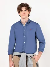 Camisa azul de hombre confeccionada en 100% algodón twill, tintada en prenda. Cuenta con corte regular fit, cuello button-down, cierre frontal de botones, manga larga con puño simple y pequeño logo rojo bordado en el pecho.