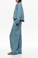 Chaqueta corta estilo boxy de denim azul claro, con cuello camisero, cierre frontal de botones metálicos a presión, mangas largas con puños abotonados y bolsillos con solapa en el pecho.