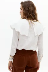 Camisa blanca de lino con cuello Peter Pan grande y ondulado, mangas largas y cierre frontal con botones de nácar. Incluye lazo decorativo de terciopelo marrón en el cuello.