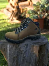 Botín de trekking color marrón con detalles en negro, confeccionado en cuero y material sintético. Presenta sistema de acordonado con ganchos metálicos, cuello acolchado y suela de goma dentada para mayor tracción.