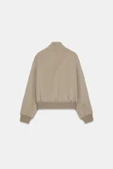 Cazadora bomber beige confeccionada en hilatura con lana, con cuello subido, manga larga y bolsillos frontales. Presenta cierre frontal con botones oscuros y puños, cuello y ruedo en tejido rib.