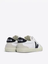 Championes Veja modelo Volley, de diseño minimalista en color blanco con detalles en negro y suela de goma en tono crema. Presentan el logo característico de la marca en el lateral y refuerzos en la puntera y el talón.