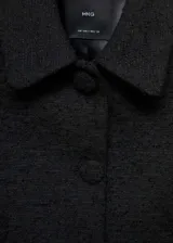 Chaqueta corta de tejido tweed en color negro, con cuello camisero, manga larga y cierre frontal con botones forrados. Presenta un diseño recto con dos bolsillos laterales.