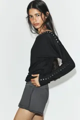 Camiseta de cuello barco y manga larga tipo murciélago, confeccionada en tejido de punto gris melange. Presenta detalle de tachas metálicas a lo largo de los hombros y las mangas.