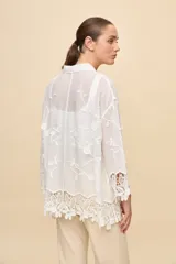 Camisa blanca de calce relajado, con cuello clásico, mangas 3/4 y cierre frontal de botones. Presenta bordados florales en relieve y detalles de crochet calado en los puños y el ruedo.