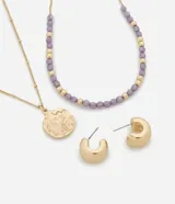 Conjunto de bisutería compuesto por dos collares y un par de aros. El primer collar presenta una cadena fina con un dije circular de textura orgánica. El segundo collar está formado por cuentas de resina en tono lila intercaladas con pequeñas esferas metálicas doradas. El set se completa con un par de aros tipo argolla gruesa con cierre de pino.