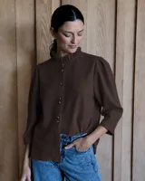 Camisa de color marrón con cuello a la base tipo mao con volados, cierre frontal con doble botonadura y mangas tres cuartos con frunces en los hombros.