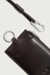 Tarjetero de piel marrón con diseño colgante para el cuello. Cuenta con ranuras para tarjetas, un bolsillo lateral con cierre de cremallera, anilla metálica y cordón largo ajustable.
