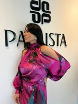 Vestido largo de satén con estampado floral en tonos fucsia, verde azulado y negro. Presenta cuello alto, aberturas en los hombros, mangas largas con puños y un lazo ajustable en la cintura.