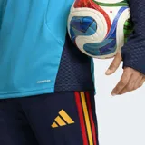 Buzo de entrenamiento Adidas Tiro España 26, color celeste con detalles en azul marino y vivos con los colores de la bandera española en los hombros. Presenta logo de Adidas en naranja y escudo de España bordado. Cuenta con cierre frontal parcial y tecnología Formotion para mayor libertad de movimiento.