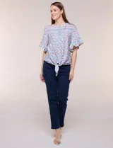 Blusa holgada de poliéster con estampado floral y geométrico en tonos celestes, azules y anaranjados sobre base blanca. Presenta cuello a la base con escote en V y botones frontales, mangas cortas amplias de estilo mariposa y un nudo en el ruedo.
