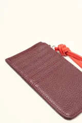 Monedero rectangular de cuero vacuno con textura granulada en color marrón topo. Cuenta con cierre superior metálico y un tirador decorativo de cuero en color naranja con un nudo. Presenta un pequeño logo grabado en el centro.