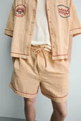 Shorts relaxed fit color beige con textura en relieve tipo jacquard, cintura elástica con cordón ajustable y detalle de bordado en contraste en el bajo.