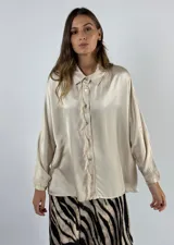 Camisa color beige de seda con cuello clásico, cierre frontal con botones y mangas largas. Presenta detalle de flecos en el frente.