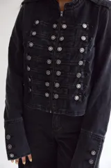 Chaqueta de estilo militar confeccionada en denim negro con efecto lavado. Presenta un diseño estructurado con cierre frontal de cremallera, cuello mao y múltiples botones metálicos decorativos dispuestos en filas horizontales. Incluye detalles de charreteras en los hombros y puños con botones.
