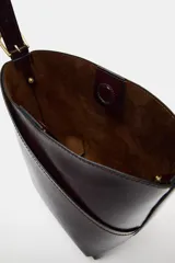 Bolso tipo saca de diseño asimétrico con acabado brillante, asa ajustable con hebilla lateral y cierre mediante botón imán.
