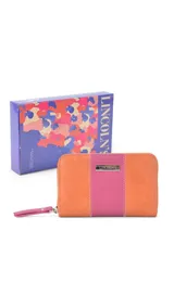 Billetera mediana de cuero sintético con diseño bicolor en naranja y fucsia. Cuenta con cierre perimetral, correa de muñeca extraíble y placa metálica decorativa con logo.