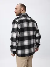 Sobrecamisa de estilo leñador con estampado de cuadros en tonos gris, negro y blanco. Presenta cierre frontal con cremallera y solapa con botones a presión, cuello clásico y bolsillos laterales.