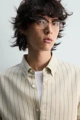 Camisa de algodón de corte relajado, con cuello solapa abotonado (button-down) y manga larga terminada en puño con botón. Presenta un patrón de rayas verticales finas en tonos beige claro y marrón/verde oliva.